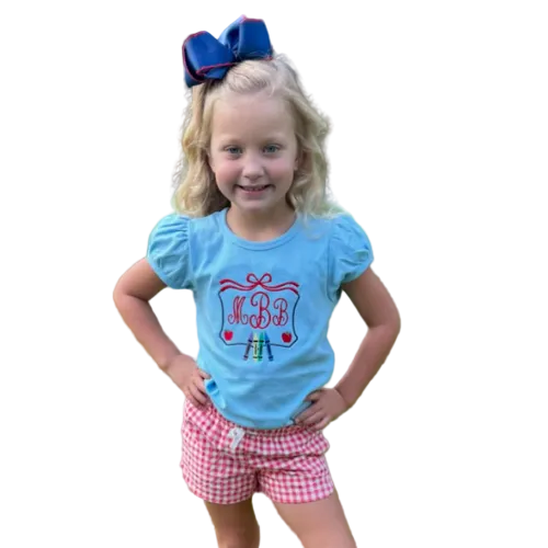 Young girl wearing a blue shirt with a design and red shorts standing on grass.
